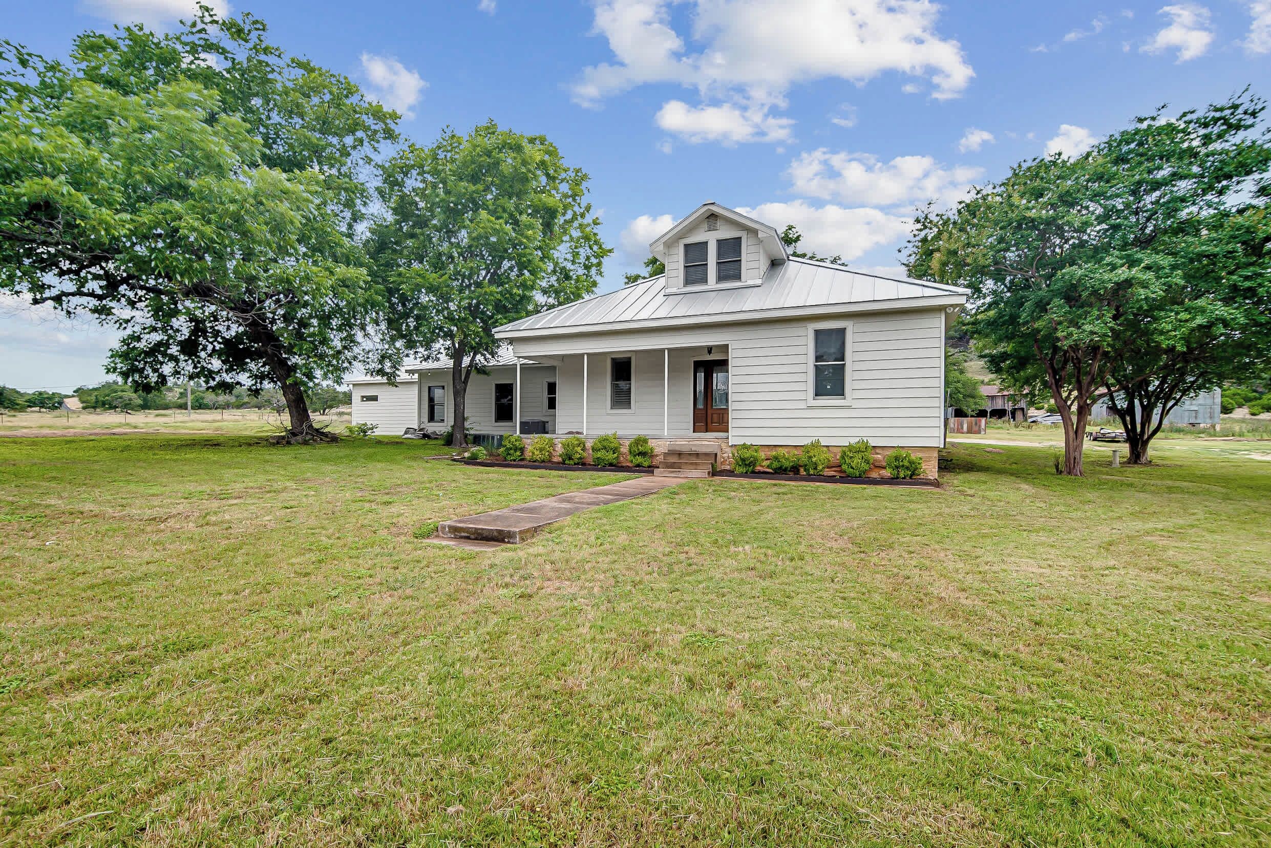 A single-story house with a front porch, a small front yard, and a large tree to the left.