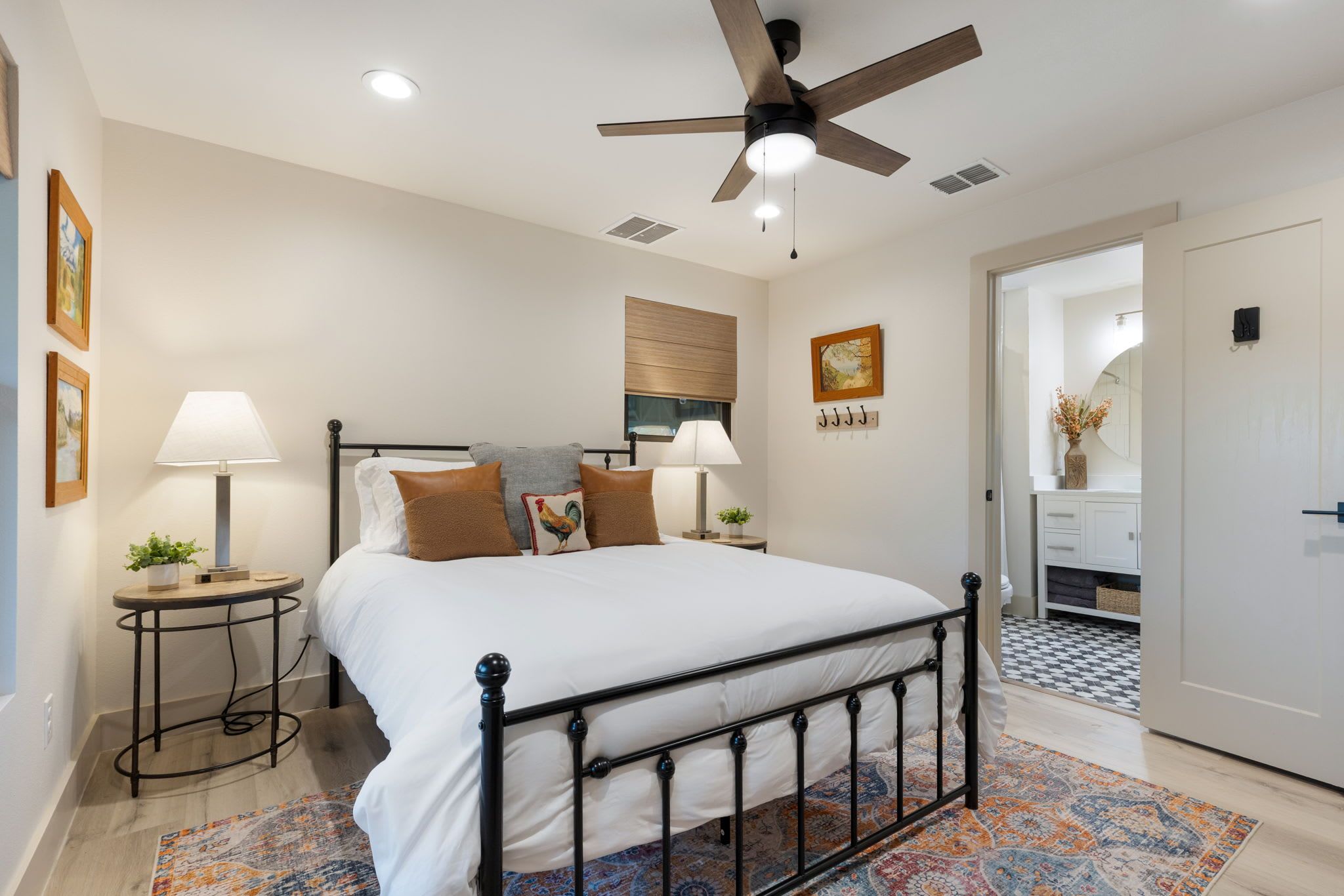 A bedroom with a bed, bedside tables, a ceiling fan, a window with blinds, and a door leading to another room.