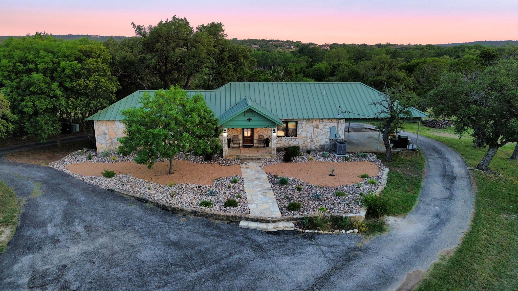 A stone house with a green roof, surrounded by a gravel driveway and a stone pathway leading to the entrance.
