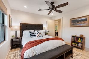 Un dormitorio con cama, ventilador de techo, mesita de noche con lámpara, estantería y ventana.