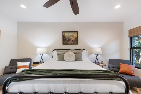 A hotel room with a large bed, two bedside tables, a ceiling fan, a painting on the wall, and a window with a view.