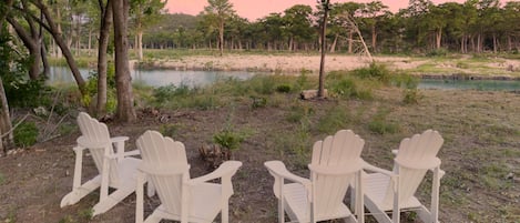 Vier witte Adirondack-stoelen zijn in een halve cirkel opgesteld, gericht op een waterlichaam, met bomen op de achtergrond.