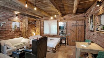 Una habitació rústica amb parets de maó al descobert, sostre de fusta, llit amb edredó blanc, una cadira de cuir, taula petita amb cadira verda i un armari de fusta.