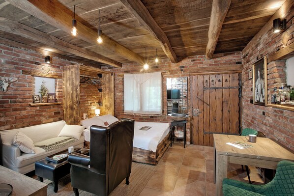 A rustic room with exposed brick walls, a wooden ceiling, a bed with a white bedspread, a leather chair, a small table with a green chair, and a wooden cabinet.
