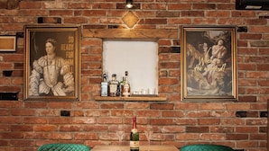 A rustic room with exposed brick walls, a wooden ceiling, and a brick fireplace. There are two green upholstered chairs, a wooden table, and a shelf with bottles and glasses. - Cozy little house near Zagreb and airport. Private garden around house. (Dugo Selo)