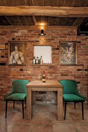 Una habitación rústica con paredes de ladrillo visto, techo de madera y chimenea de ladrillo. Hay dos sillones tapizados en verde, una mesa de madera y una estantería con botellas y copas.