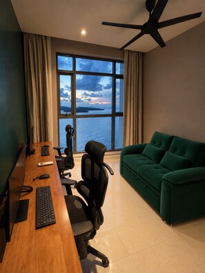 A room with a desk, office chair, and a green sofa. There is a window with a view of the sea and a ceiling fan. - Seafront - Spacious 4-bedroom apartment in charming Langkawi with AC (Langkawi)