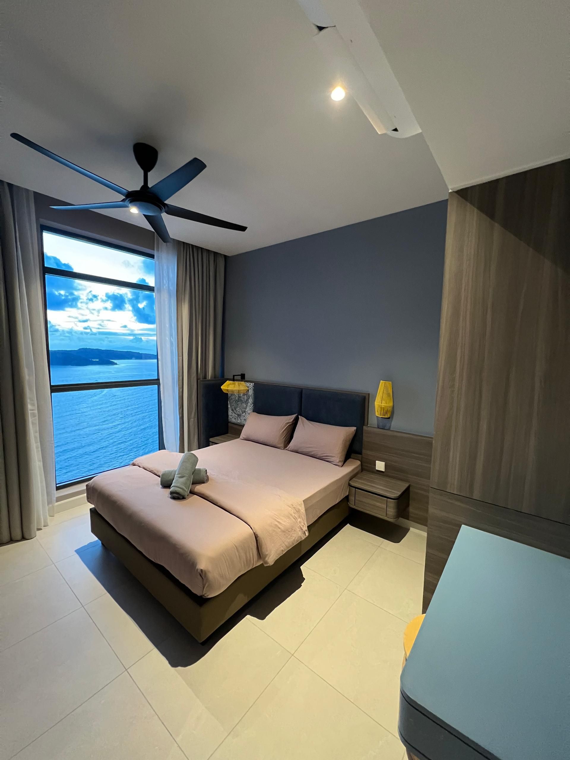 A modern bedroom with a large bed, a ceiling fan, a nightstand, and a view of the sea.