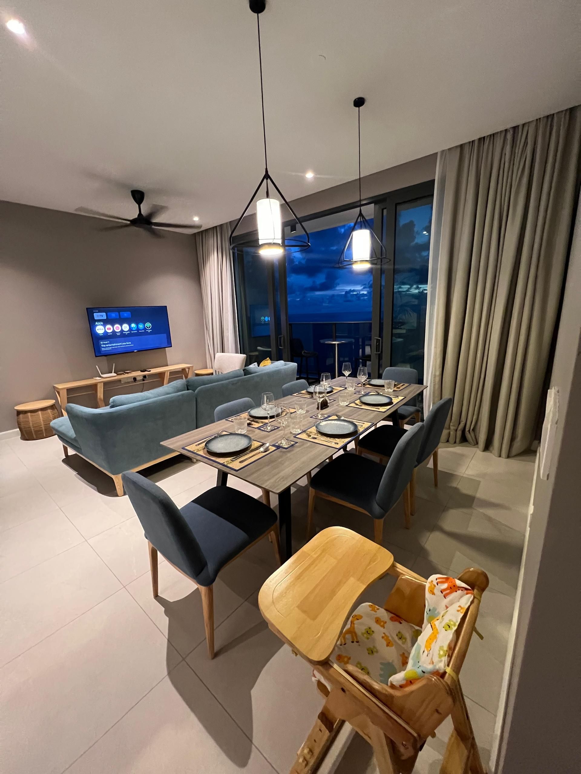 A modern dining area with a wooden table set for four, blue upholstered chairs, and a television displaying a menu.