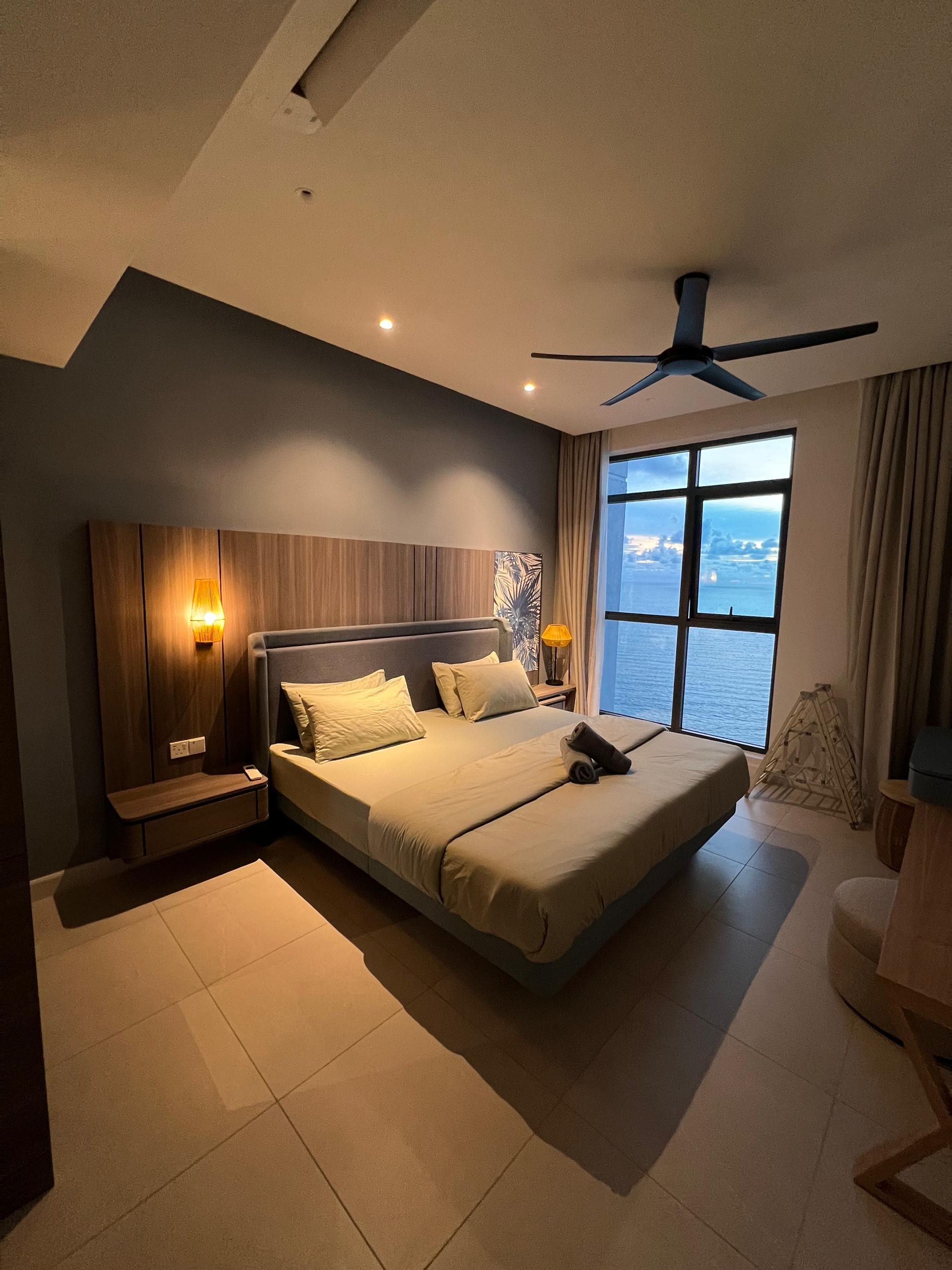 A modern bedroom with a large bed, a ceiling fan, and a view of the sea.