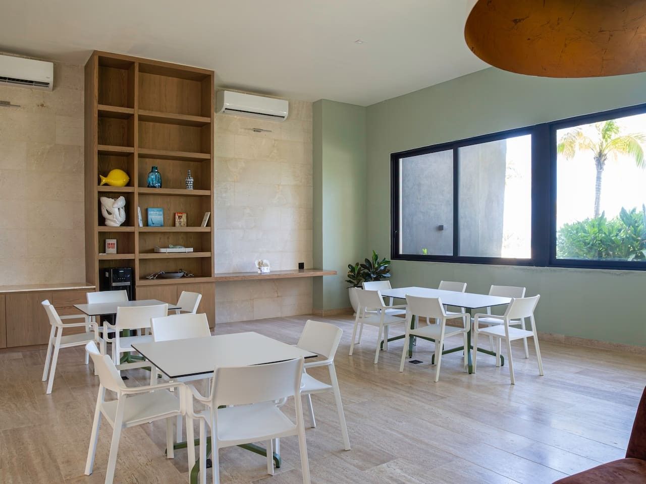 A room with a wooden bookshelf, white tables and chairs, a large window with a view of palm trees, and an air conditioning unit on the wall.