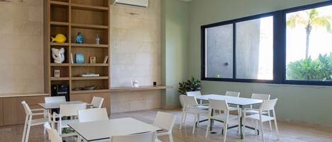 A room with a wooden bookshelf, white tables and chairs, a large window with a view of palm trees, and an air conditioning unit on the wall.