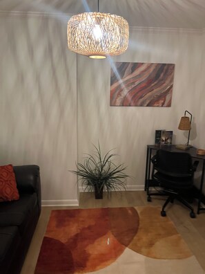 Una habitació amb un sofà de cuir negre, una catifes marró i taronja, una planta en test, un escriptori amb una làmpada i un quadre a la paret.