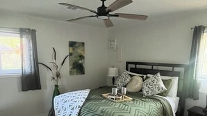 Quarto com cama, ventilador de teto, mesa de cabeceira, luminária, janela com cortinas e abajur.