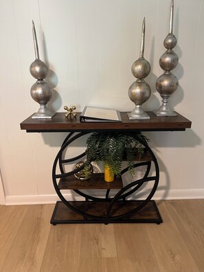 Una consola de madera con base metálica, dos candelabros decorativos, una planta pequeña y un objeto esférico.