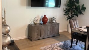 Sala de estar com televisão de tela plana na parede, aparador de madeira com objetos decorativos, mesa de jantar com cadeiras e uma planta grande em vaso.