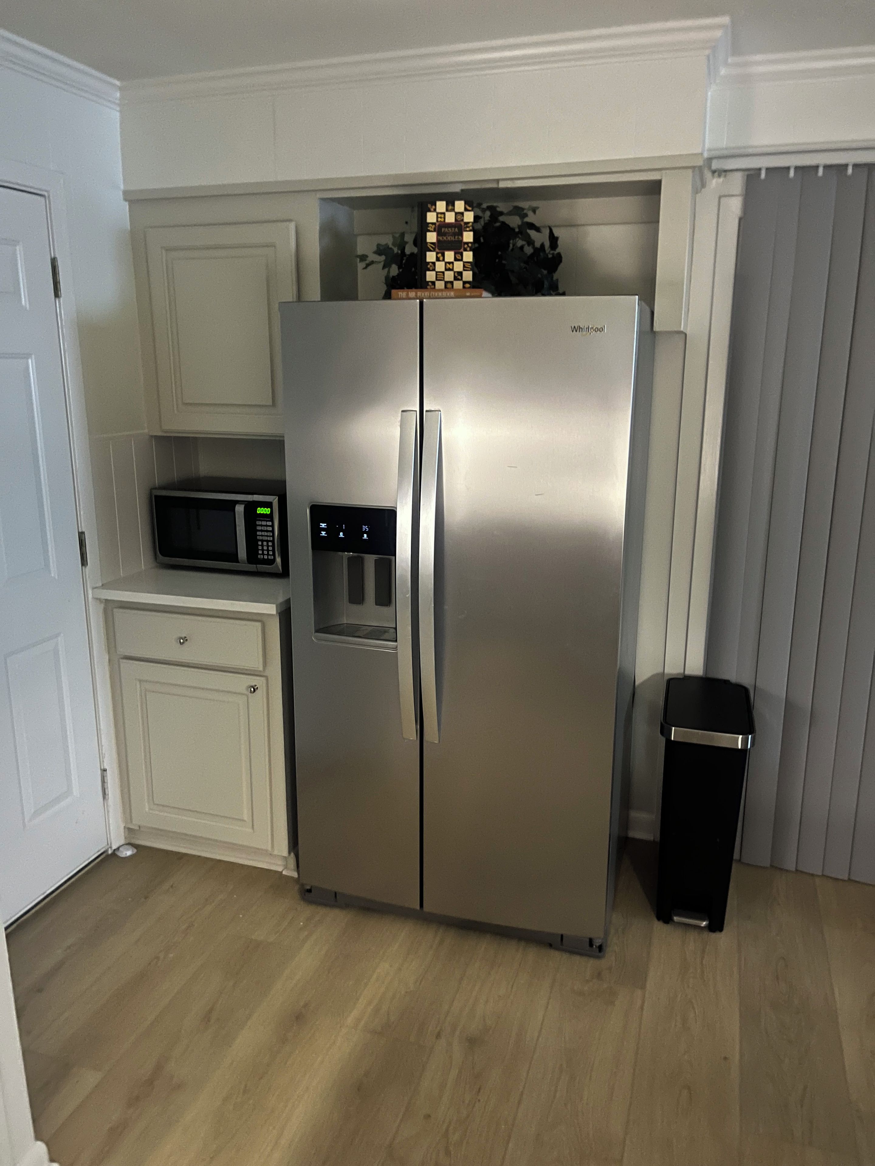 A stainless steel refrigerator with a digital display, a microwave, and a trash bin in a kitchen with white cabinetry and a wooden floor.