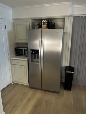 A stainless steel refrigerator with a digital display, a microwave, and a trash bin in a kitchen with white cabinetry and a wooden floor.