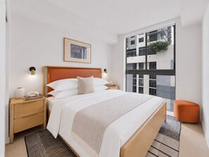 Ein modernes Schlafzimmer mit einem großen Bett, einem Nachttisch, einer Nachttischlampe, einem gerahmten Bild an der Wand und Blick ins Freie durch ein großes Fenster.