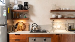 Une cuisine compacte équipée d’un réfrigérateur, d’une cuisinière, d’une bouilloire, d’une machine à café et de divers ustensiles rangés sur des étagères en bois.