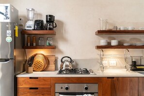 Eine kleine Küche mit Kühlschrank, Herd, Wasserkocher, Kaffeemaschine und verschiedenen Küchenutensilien auf Holzregalen.