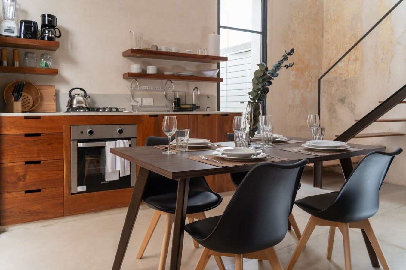 A modern kitchen with wooden cabinets, a dining table set for four, and a staircase in the background.