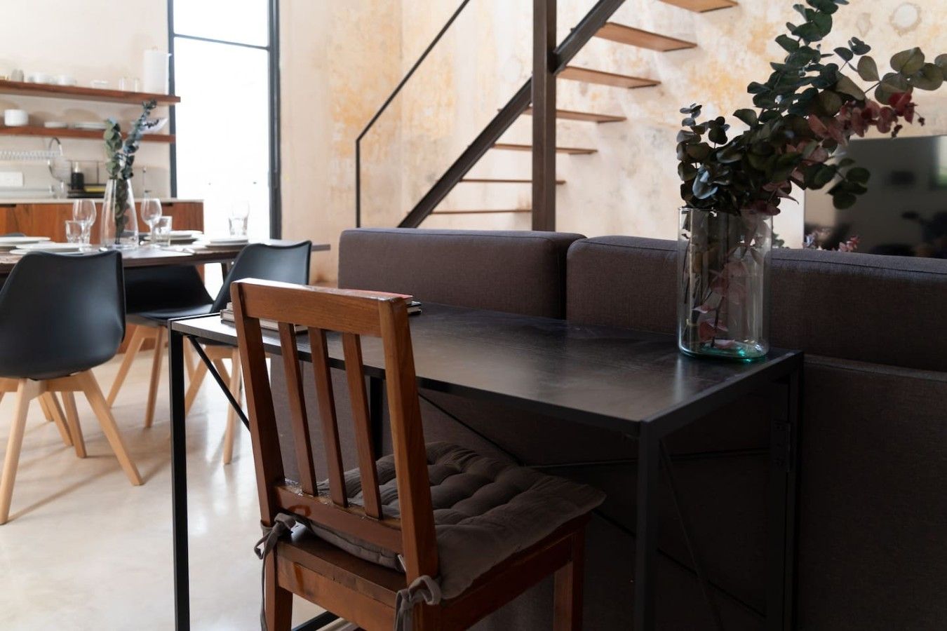 A modern dining area with a wooden chair, a glass vase with a plant, and a sofa.