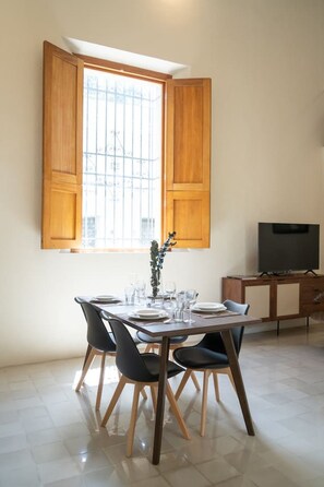 Een eetgedeelte met een tafel voor vier personen, een televisie op een standaard en een raam met houten luiken.
