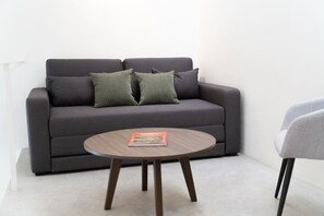 Een grijze bank met twee groene kussens en een ronde houten salontafel met een tijdschrift.