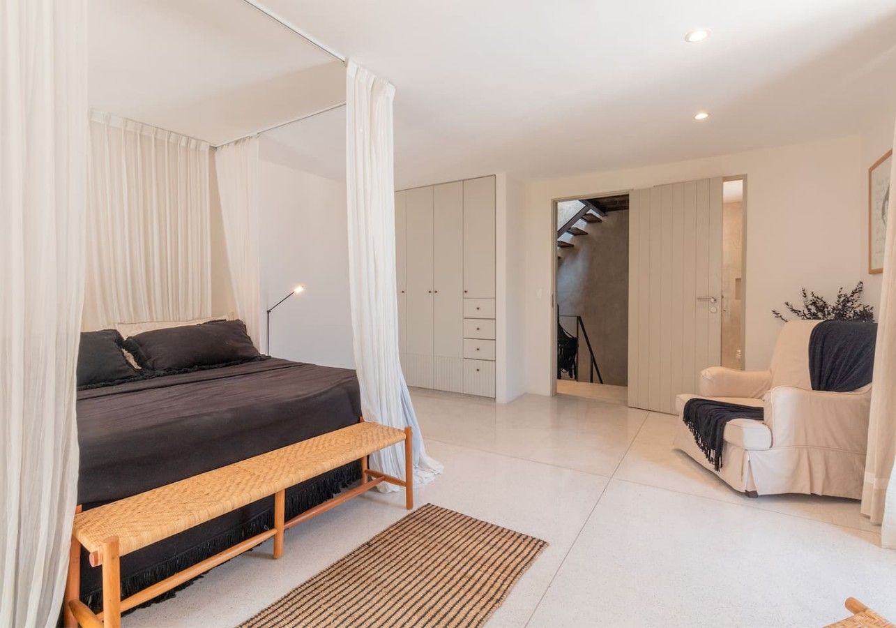 Une chambre à coucher comprenant un lit, une table de chevet, un banc et une chaise. On y trouve également une armoire et une porte qui donne sur une autre pièce.