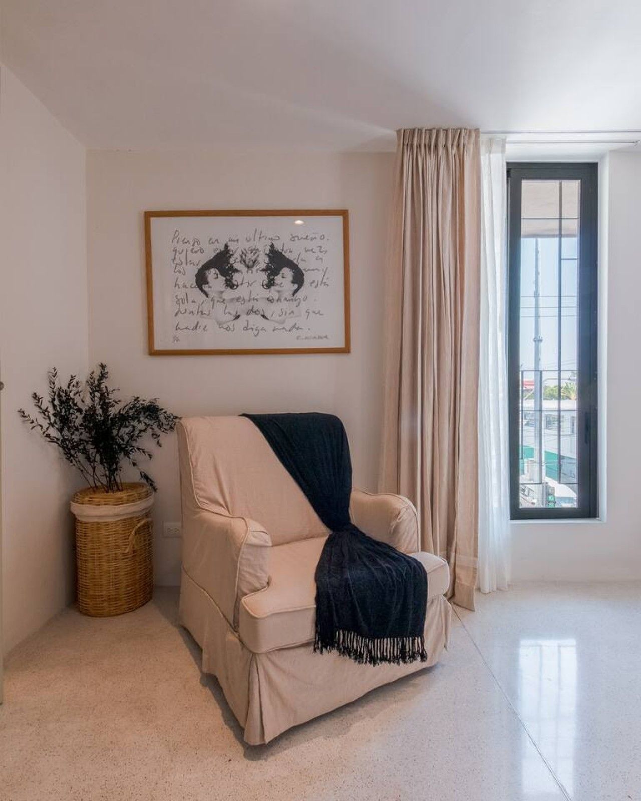 Una habitación con un sofá color beige, una obra de arte enmarcada en la pared, una planta en maceta y una ventana con cortinas.