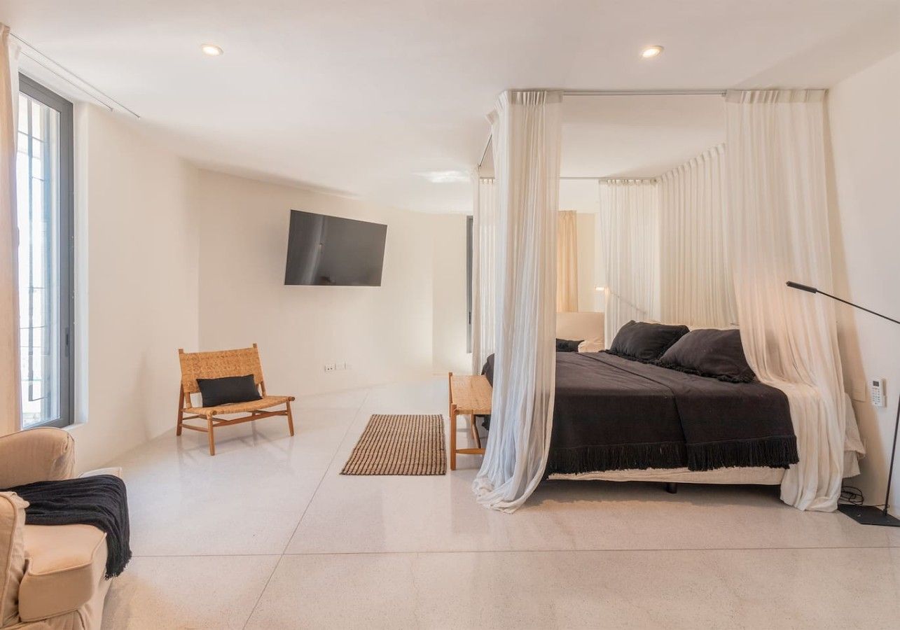 A bedroom with a canopy bed, a flat-screen TV on the wall, and a seating area with a chair and a sofa.