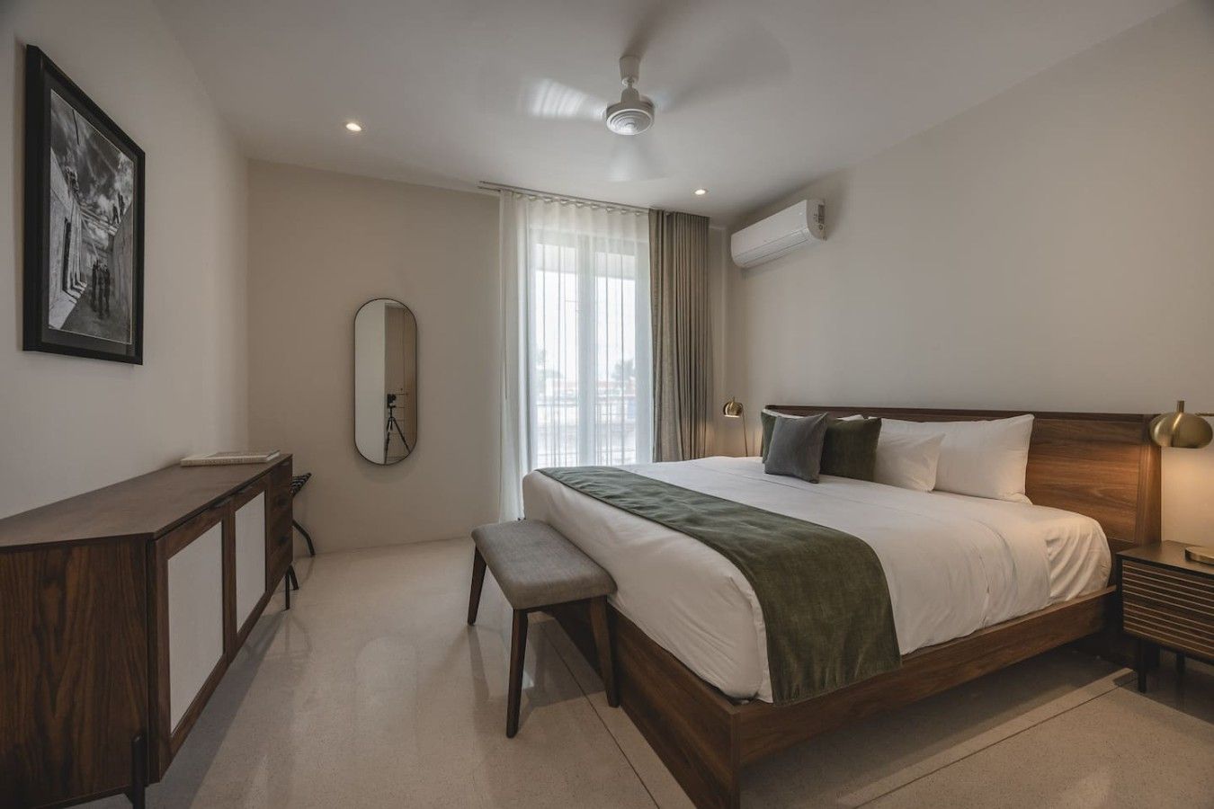 Una habitación de hotel con cama, mesita de noche, banco, espejo y unidad de aire acondicionado.