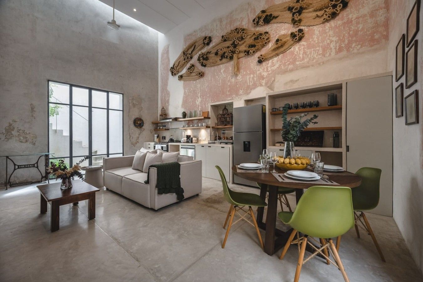 A spacious living area with a sofa, dining table, and chairs, a kitchen in the background, and a large window providing natural light.