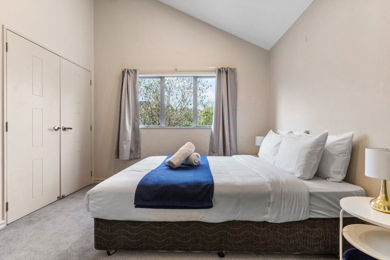 Een hotelkamer met een bed, een raam met gordijnen, een nachtkastje met een lamp en een deur.