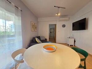 A modern living room with a round table, a sofa, a TV, and a window with curtains. - Quiet 1BR + Pool, 300m to beach, 100m to Supermarket, Walk to Dining (Limassol)