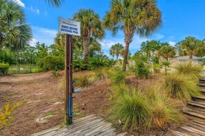 A sign indicating to rinse before entering a pool, surrounded by a natural landscape with palm trees and a wooden deck. - Bikini Bottom · New Listing! Steps to Beach, Gulf-View Porches & Pool (Gulf Shores)