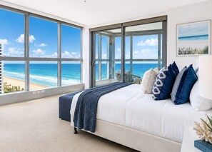 A bedroom with a large window overlooking a beach, a bed with blue and white bedding, and a view of the ocean. - Harmony Oceanview 2BR Suite | Jewel + Parking (Surfers Paradise)