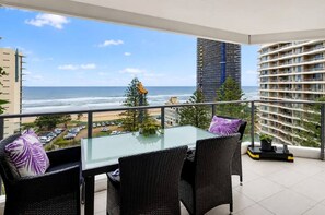 A balcony with a glass table and wicker chairs, overlooking a beach and cityscape. - Beachfront Surfers Paradise Apartment - Stunning Ocean Views (Surfers Paradise)