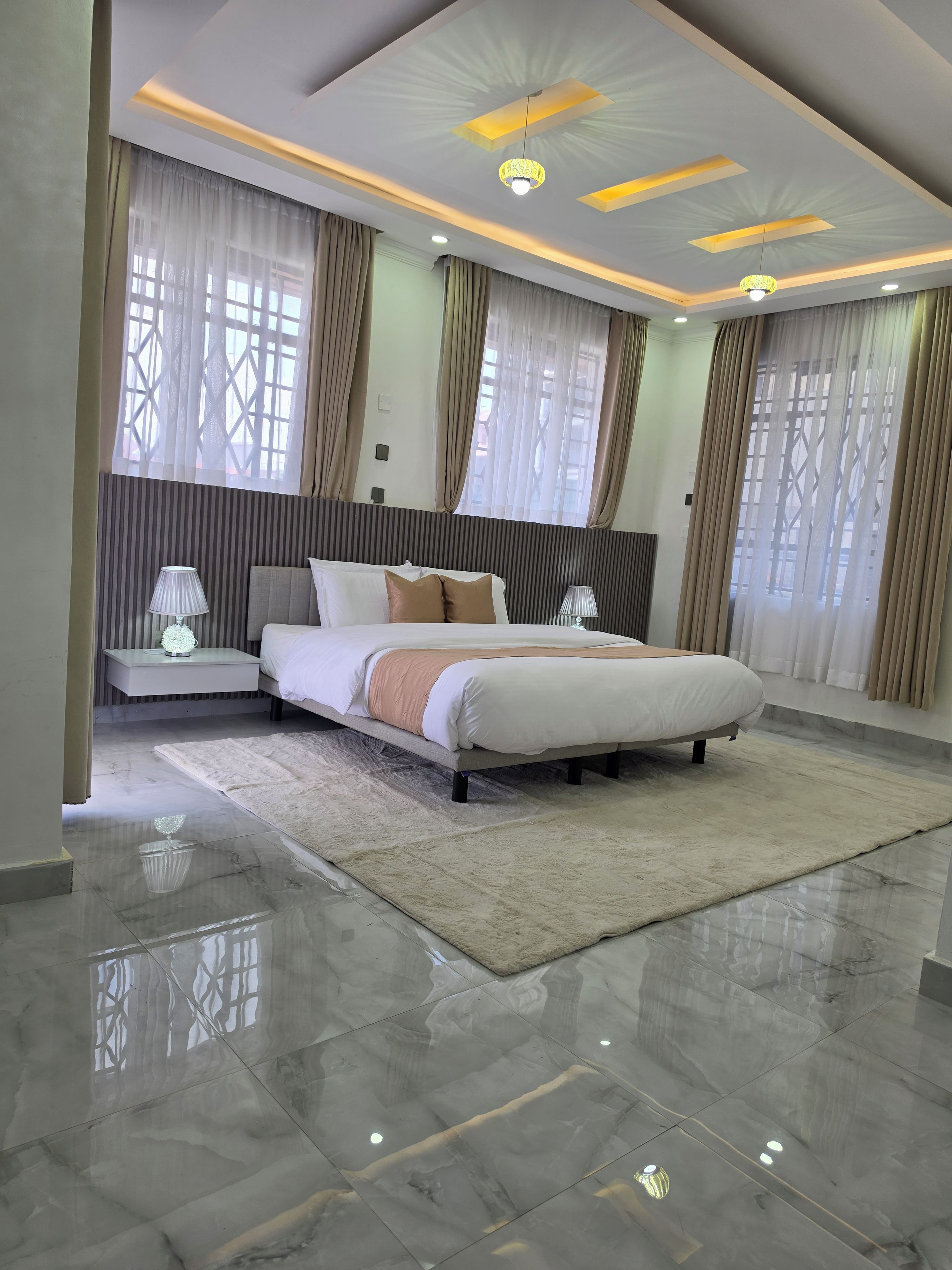 A modern bedroom with a large bed, bedside lamps, a decorative ceiling, and a view through the window.