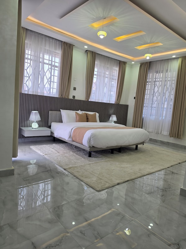 A modern bedroom with a large bed, bedside lamps, a decorative ceiling, and a view through the window.