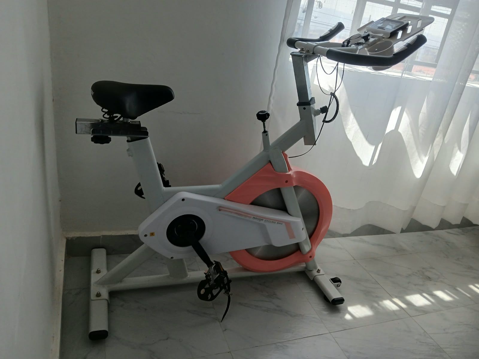 A stationary exercise bike with a digital display, positioned in a room with a tiled floor and a window with sheer curtains.