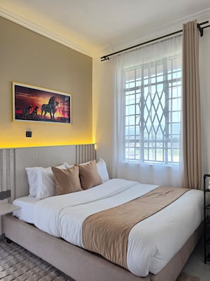 A hotel room with a large bed, a headboard, a window with curtains, and a wall-mounted painting.