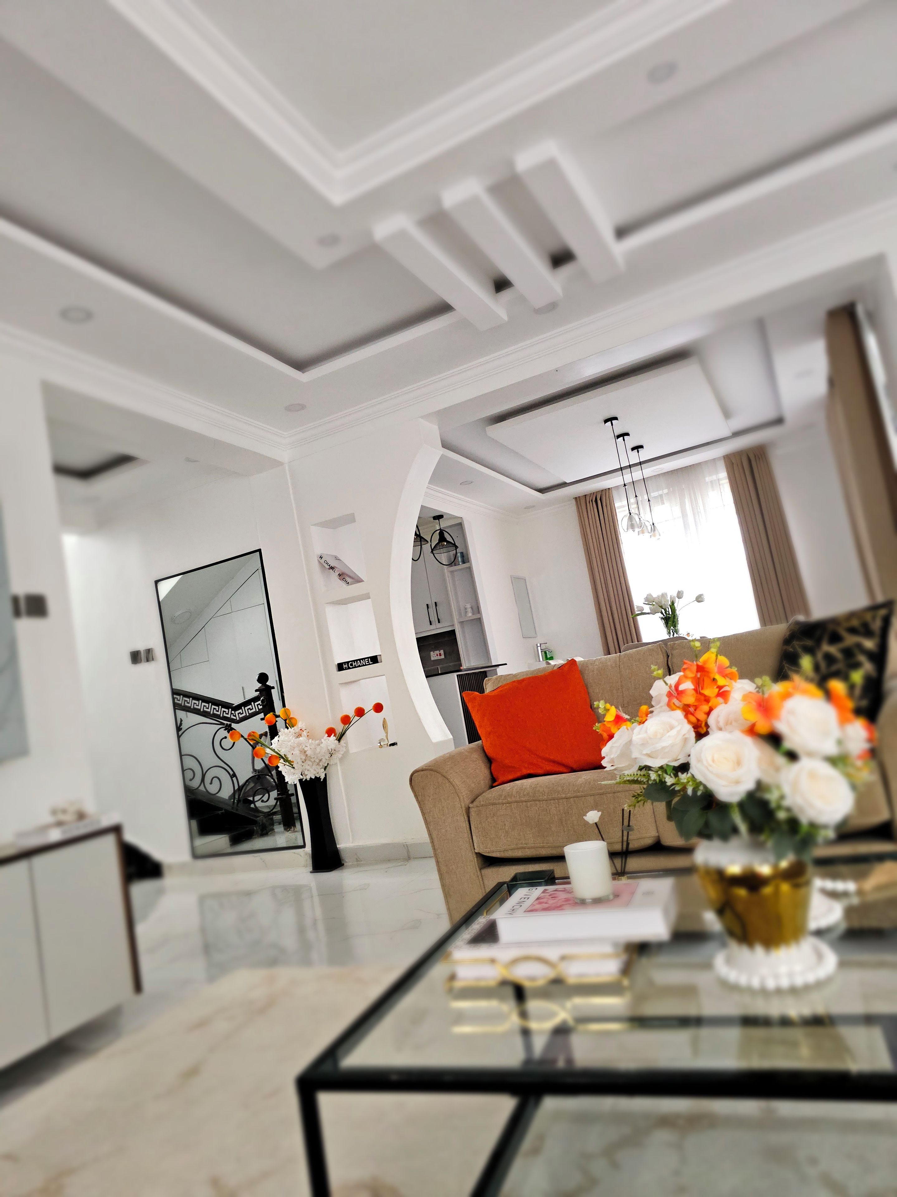 A modern living room with a glass coffee table, a beige sofa with a red cushion, and a vase of flowers.