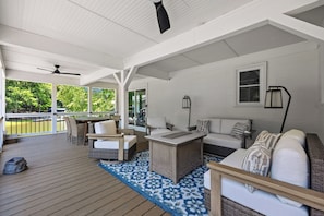 A covered outdoor seating area with a wooden deck, white walls, and a view of greenery.