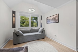 A modern living room with a grey sofa, a large window with a view of trees, a chandelier, and a framed landscape painting. - Amazing One of a kind 3 Bedroom Lakeside Chalet with Optional Boat Rental (Rock Hill)