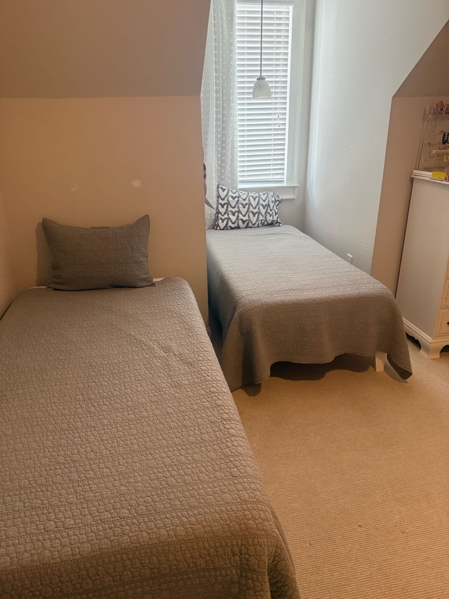A bedroom with two beds, a window with blinds, a hanging light, and a dresser.