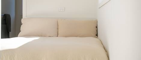 A bed with a beige bedspread and two pillows, a power outlet on the wall, and a window on the right side.