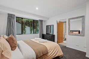 A modern bedroom with a large bed, a dresser, a mirror, and a view of greenery outside. - Stunning Lake Views - Queenstown (Queenstown)