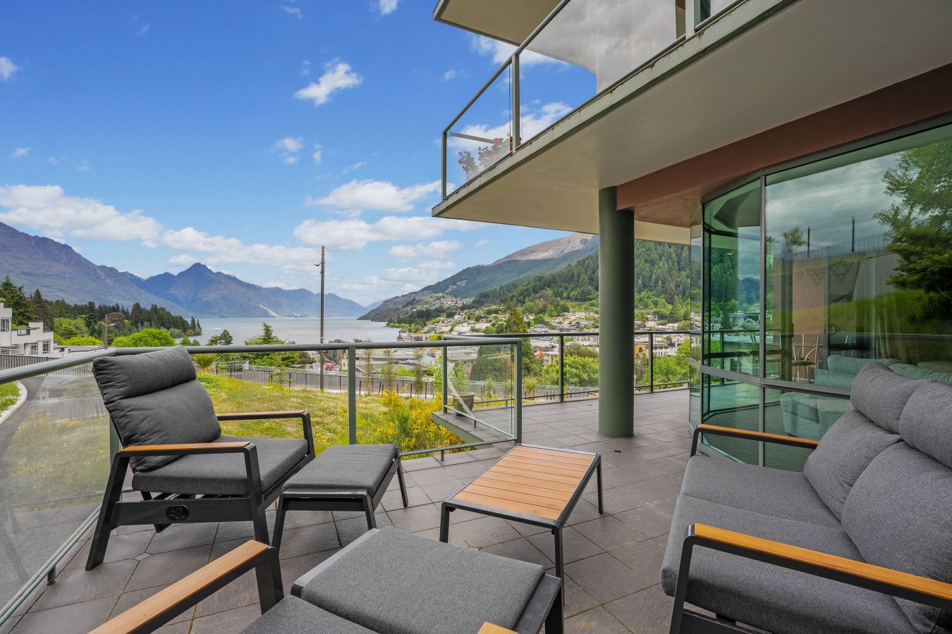 A balcony with outdoor seating, a glass railing, and a view of mountains and a lake.
